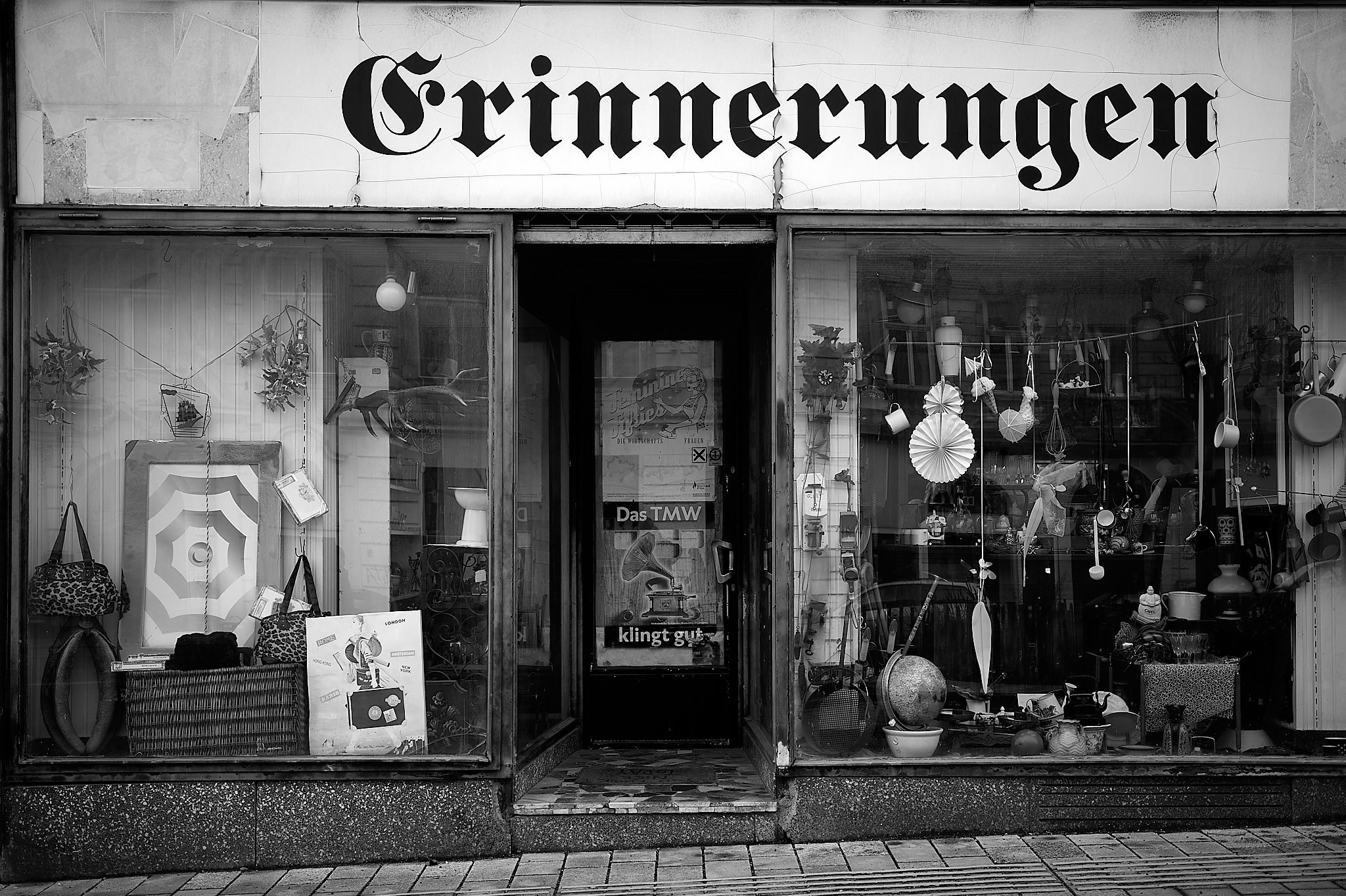 A black and white image of a quaint storefront with the word "Erinnerungen" on the sign above, suggesting a German-speaking location. The window displays various items including bags, sculptures, wall decorations, and small lighting fixtures. A reflection on the door shows the street opposite, hinting at an urban setting.
