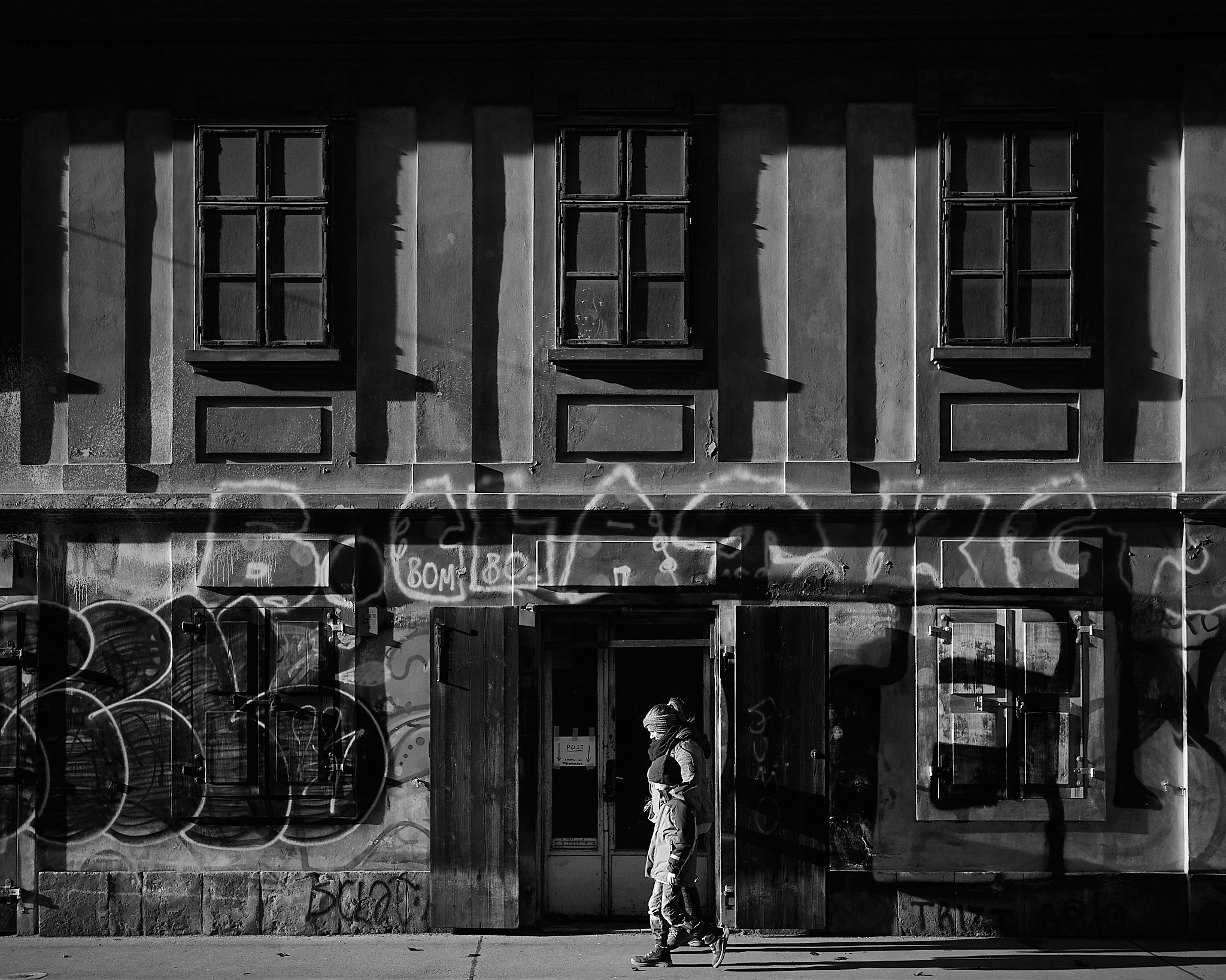 A black and white photograph depicts an adult and a child walking side by side past a building with a textured facade, adorned with graffiti and partially illuminated by sunlight casting strong shadows from window shutters.
